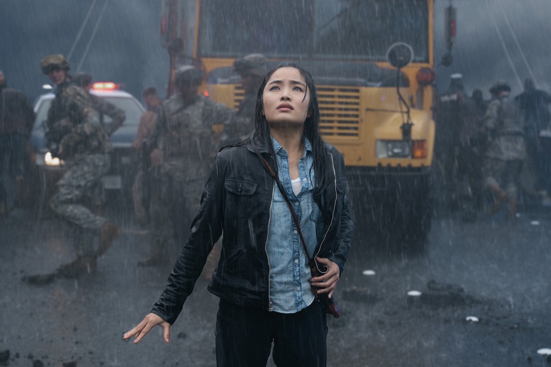 Anna Sawai, as Cate, looks upward in shock while standing in heavy rain near soldiers.