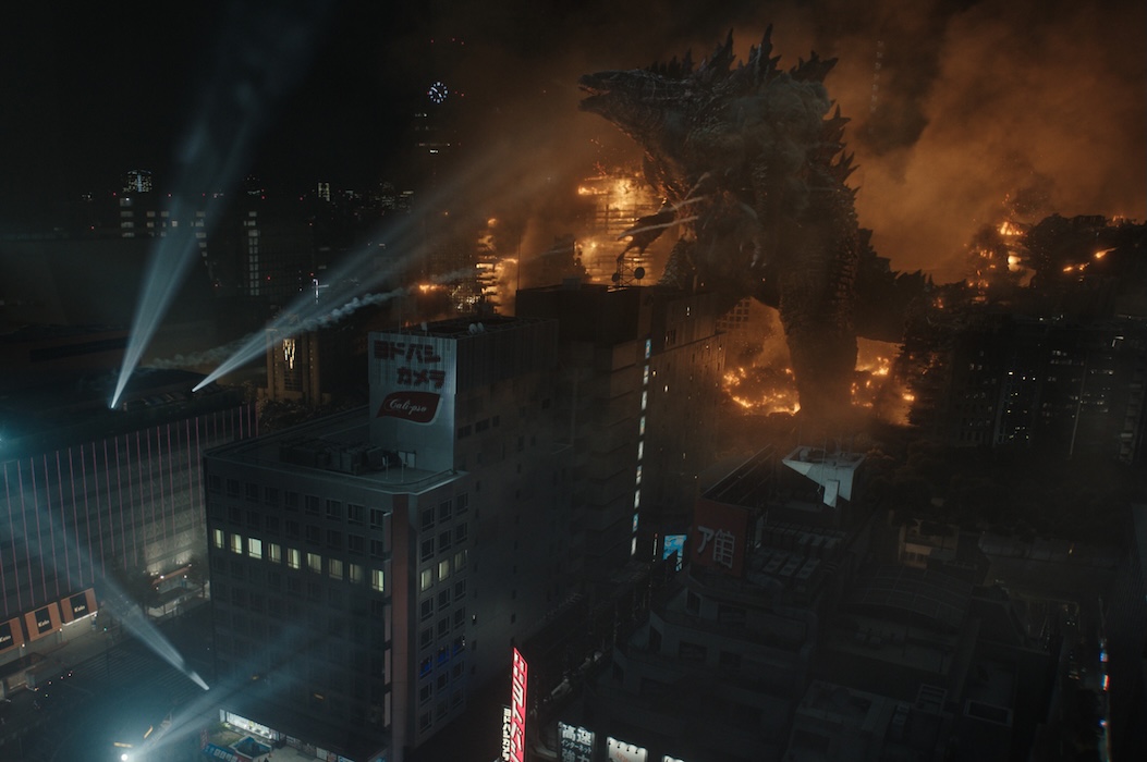 Godzilla towers over a city at night, surrounded by smoke, fire, and destruction.