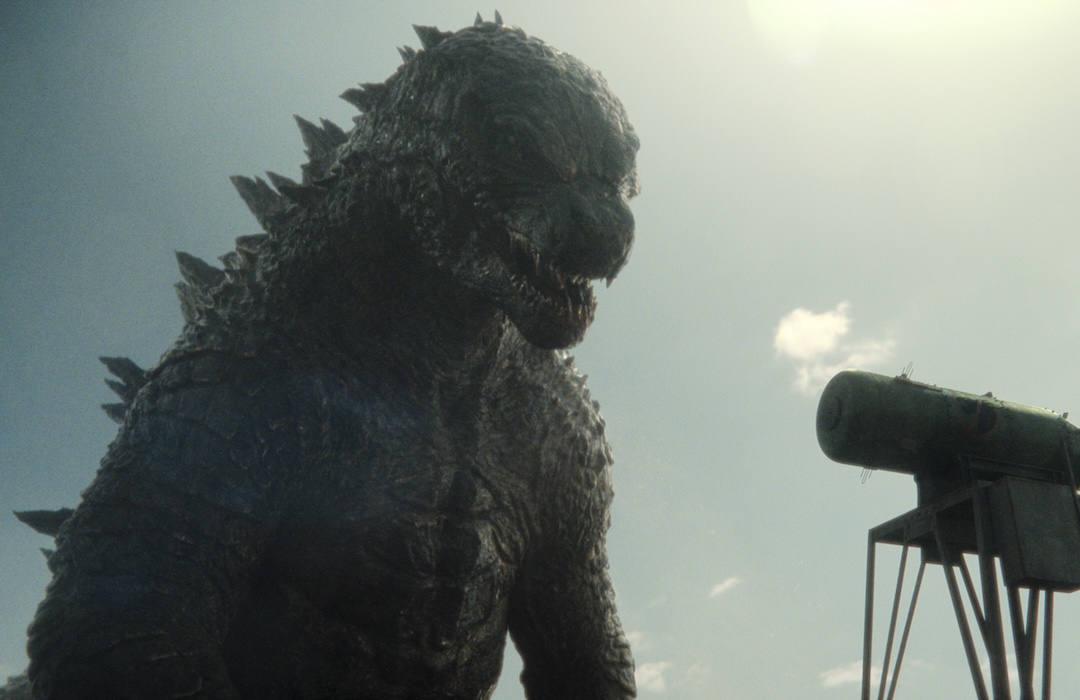 Godzilla stands tall under a bright sun, looming menacingly over a green industrial tank.