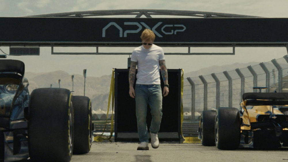 Ed Sheeran walks towards the camera, head looking down, between two Formula 1 cars on a racing track.