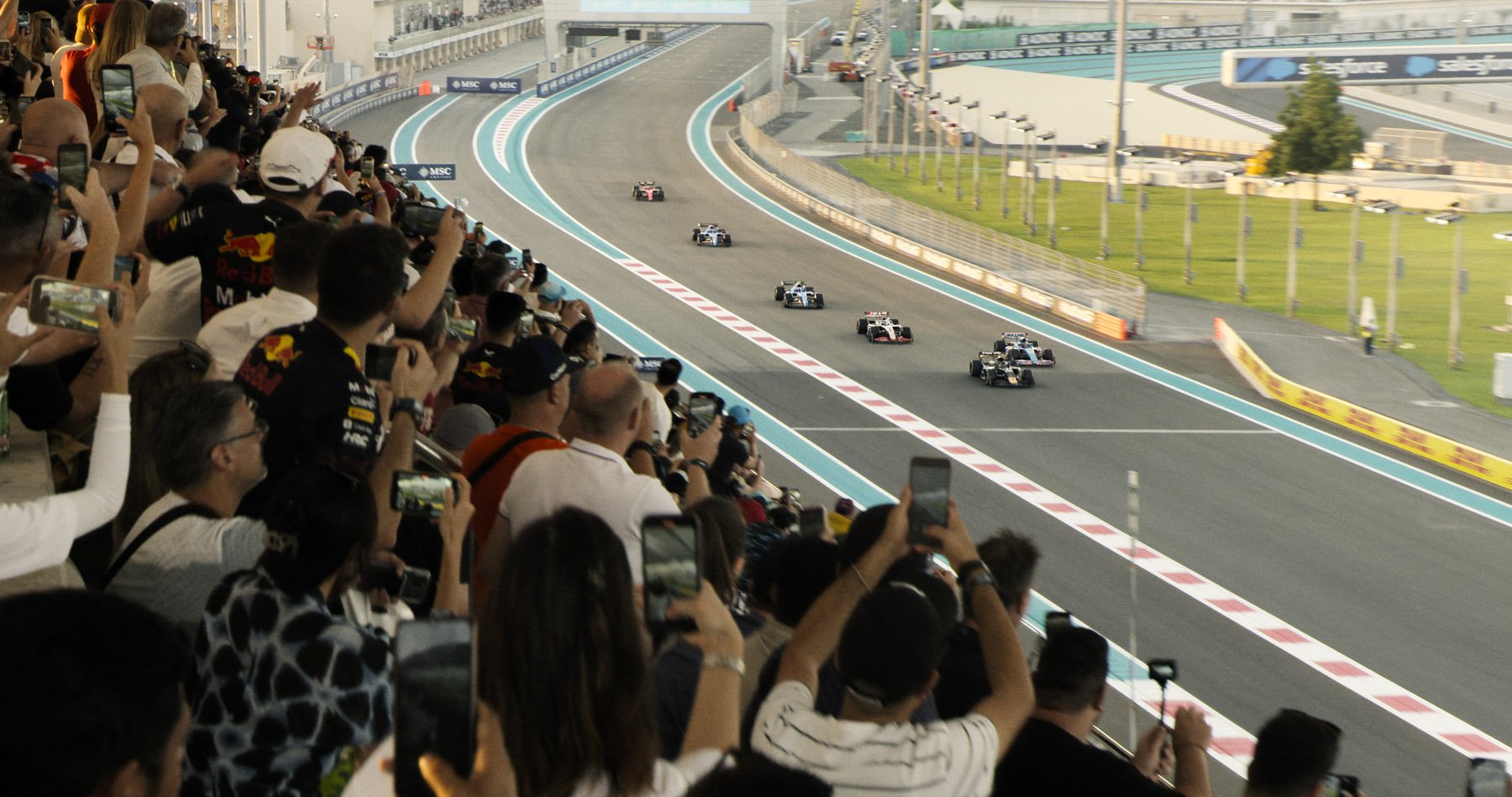 Spectators in the stands at a Formula 1 race stand up in excitement as F1 cars speed past them